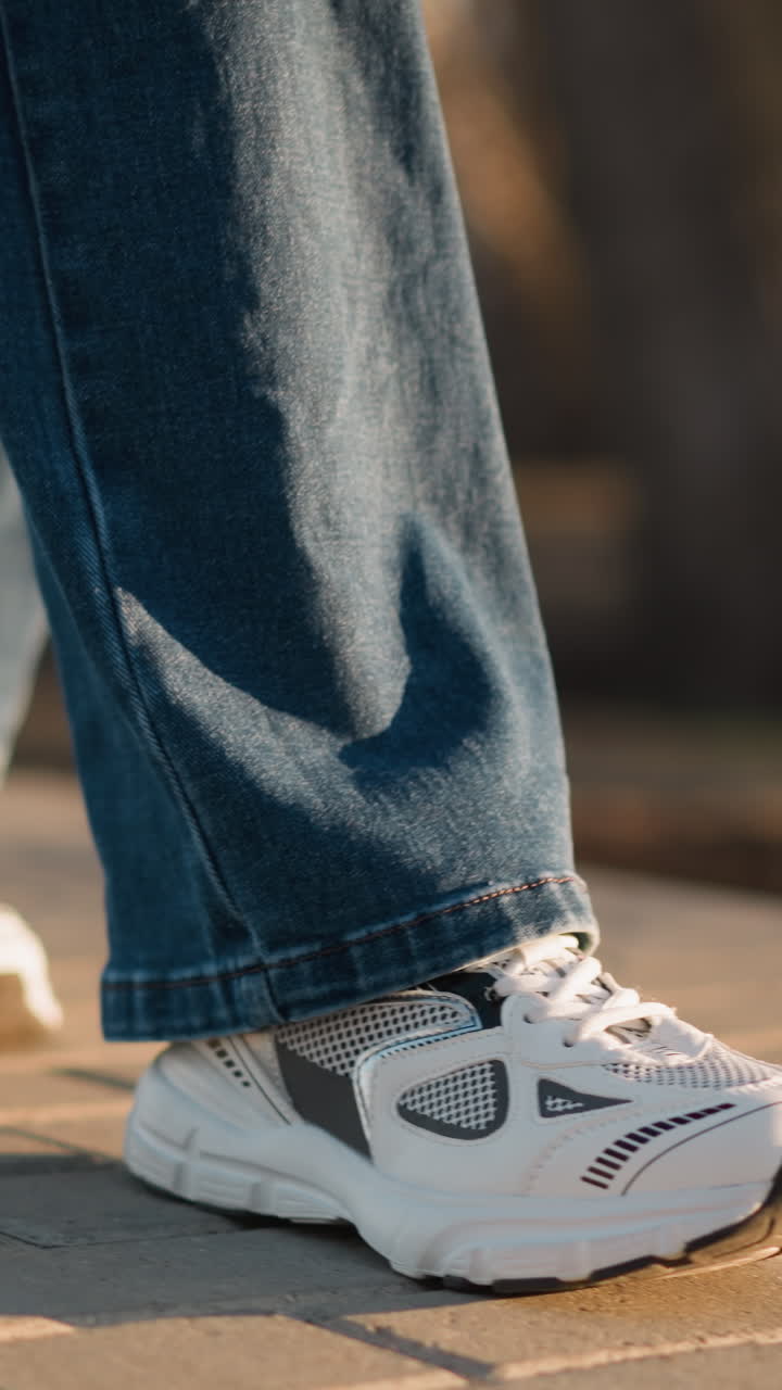 A close-up view of a person wearing jeans and white shoes, walking slowly on a paved path in a park. calm and deliberate pace of the steps, conveying a sense of sadness and introspection