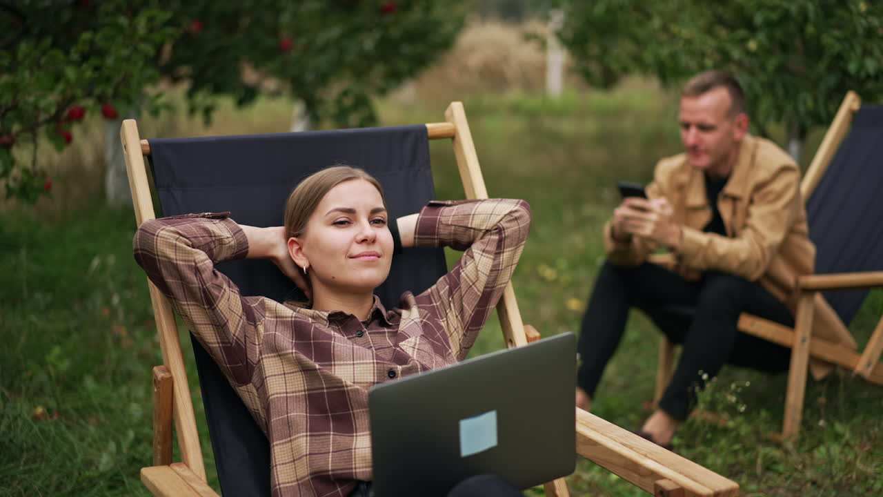 Man and woman sit in garden chairs in the nature. Lady is having a break from work on laptop and male using smartphone. Apple orchard backdrop.
