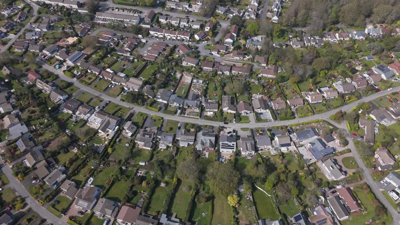 Aerial top-down of concentric suburban streets where detached homes with gardens and light roofs form elegant arcs amid lawns, hedges and surrounding woodland near the coast