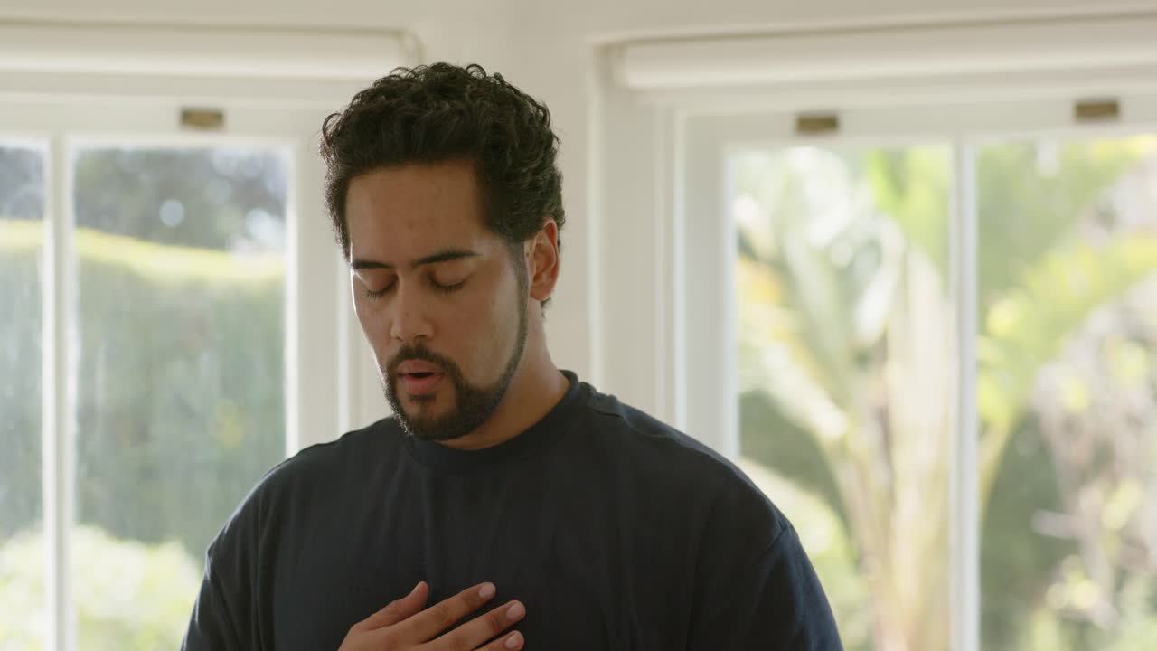 Uomo afroamericano di mezza età che si tocca il petto e l'addome, riscaldando la voce vicino alla finestra.