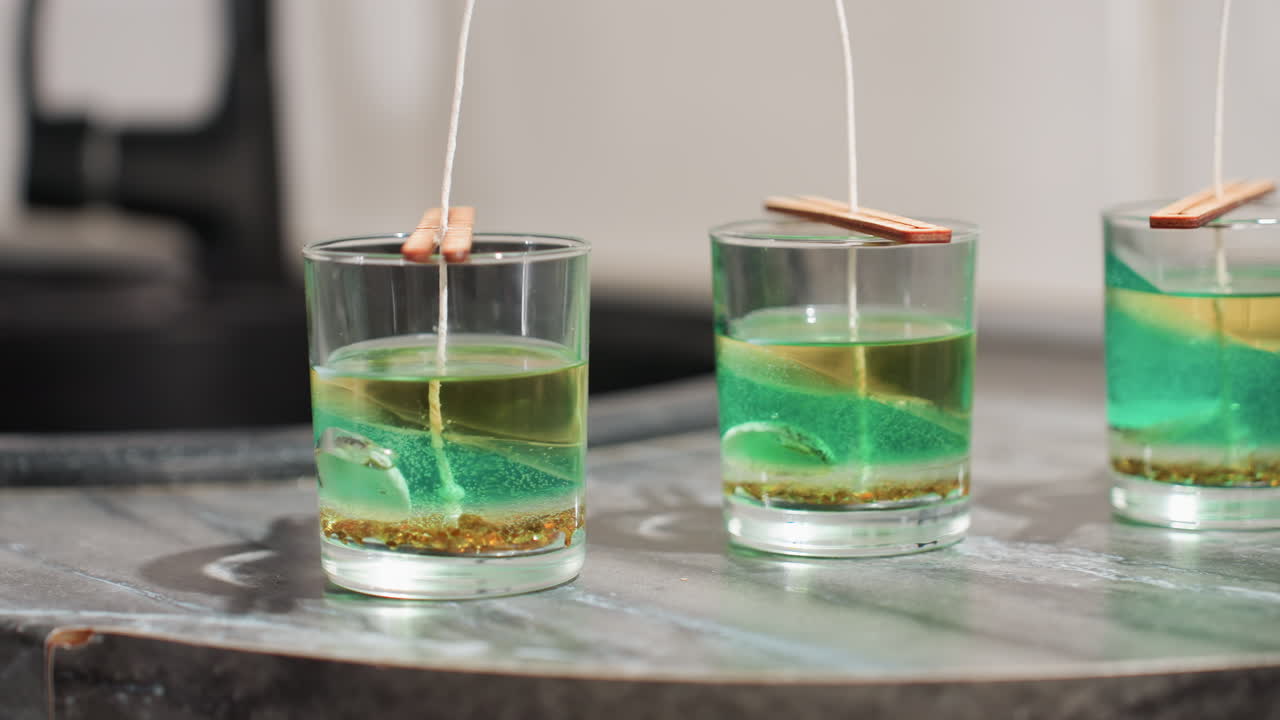Round view of transparent molds filled with melted wax as wicks held firm by sticks rest on top of each container, showcasing early stage of candle-making process in well-lit kitchen