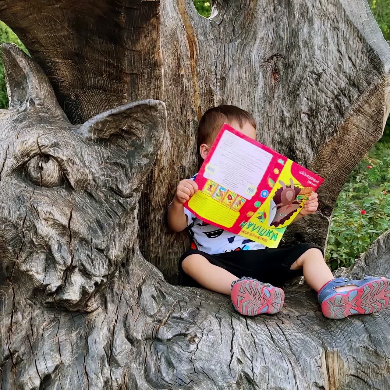 Bambino caucasico sorridente seduto sulla scultura in legno di un gatto. Bambino felice con un libro all'aperto.