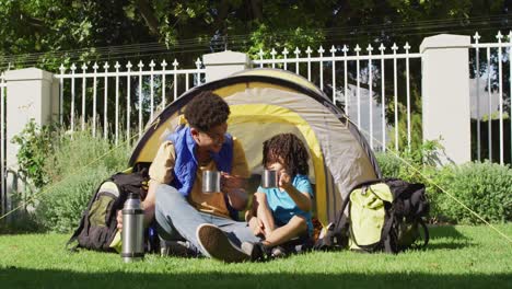 Happy-biracial-man-and-his-son-drinking-from-thermos-flask-in-garden