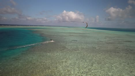 Kitesurfista-Deslizándose-Sobre-Aguas-Cristalinas-De-Color-Turquesa,-Seguimiento-De-Drones,-Día-Soleado,-Ambiente-Tropical,-Vista-Aérea