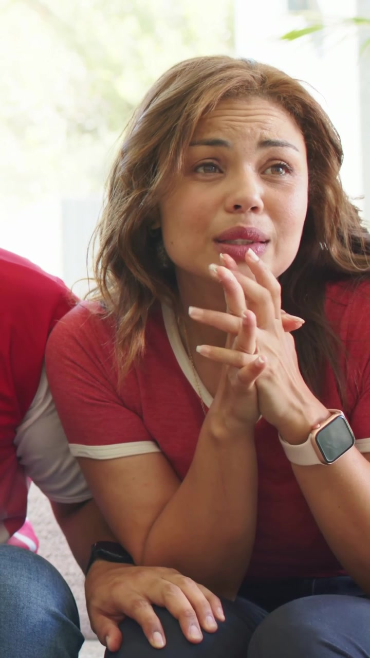 Video vertical: Mujer con top rojo reaccionando a su pareja, con las manos entrelazadas en el sofá, mostrando un reloj inteligente.