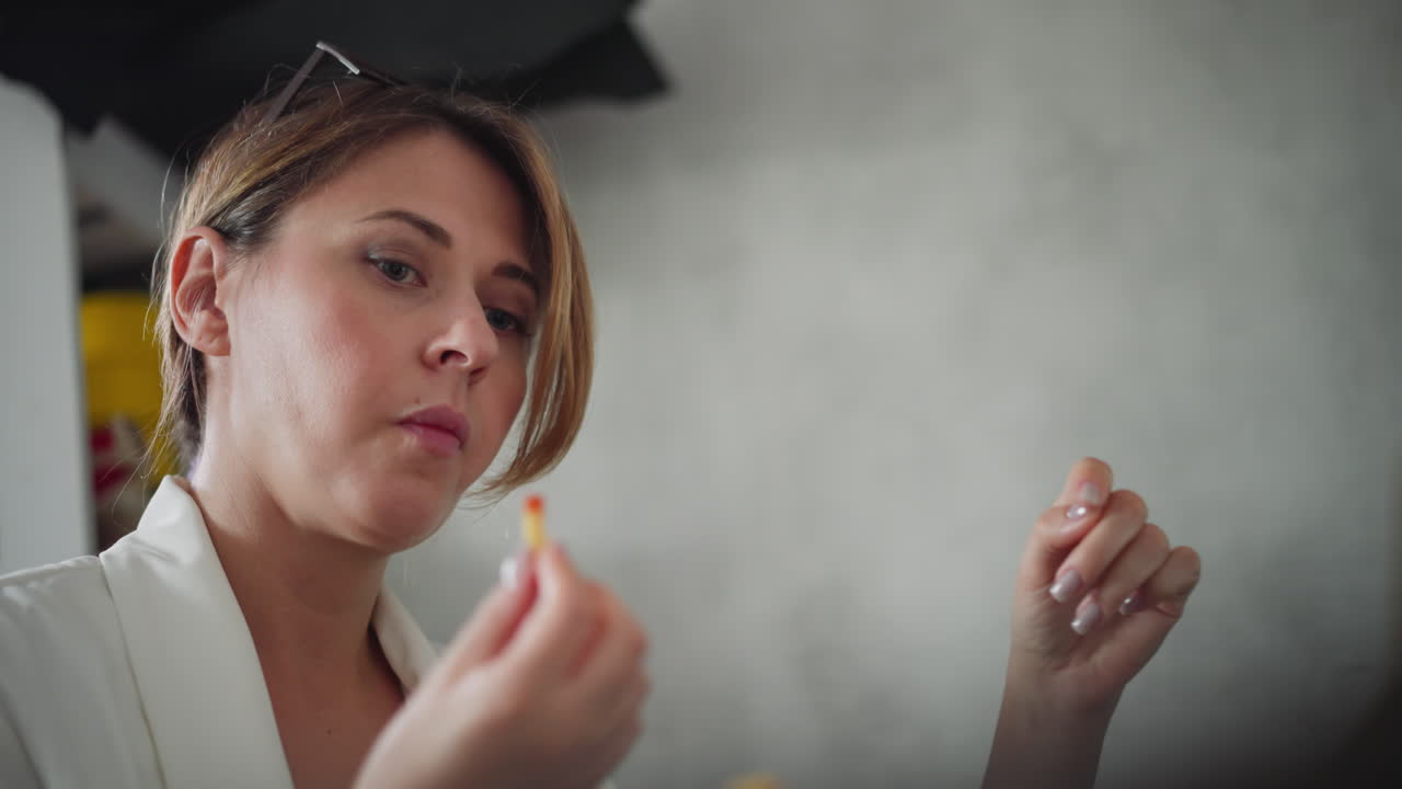 Side view of female doctor wearing white coat eating fries dipped into red sauce focusing back on her work with serious expression in simple office environment with soft lighting