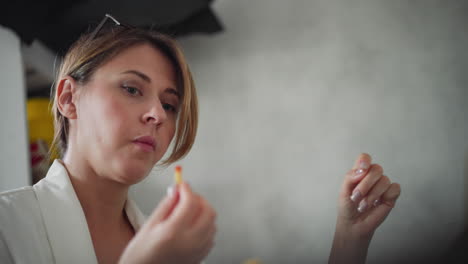 side view of female doctor wearing white coat eating fries dipped into red sauce focusing back on her work with serious expression in simple office environment with soft lighting