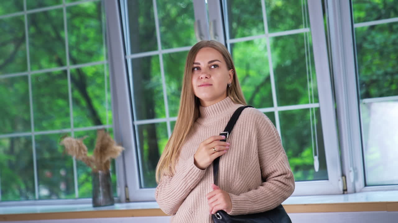 Young blonde woman in beige sweater standing indoors at the window. Beautiful lady holding a black bag on her shoulder.