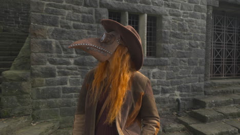 redhead wearing plague mask and cowboy hat standing among stone steps and stone buildings on bright autumn day