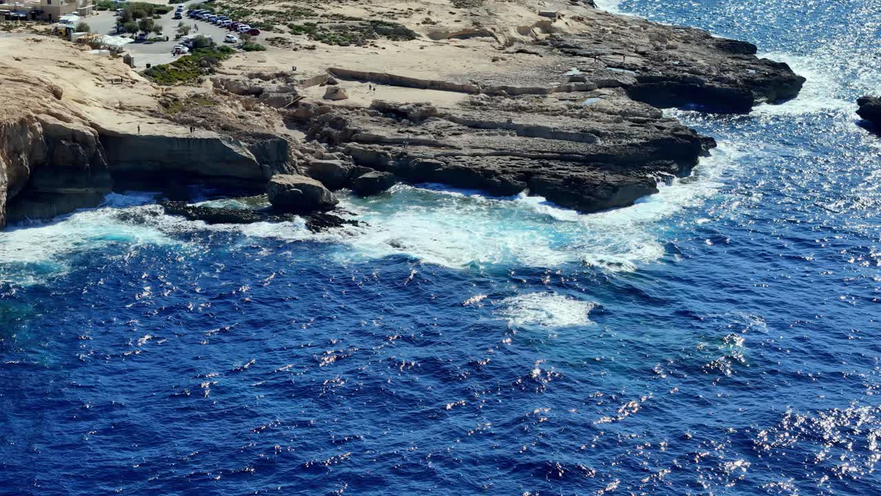 Prospettiva aerea da angolazione elevata che cattura potenti onde bianche che si infrangono sulle frastagliate scogliere calcaree di Gozo, Malta, sotto un sole estivo splendente, mettendo in mostra le acque blu intenso del Mediterraneo e il terreno roccioso