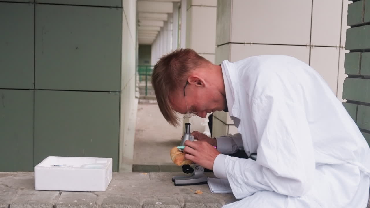 Side view of botany student in white coat carefully examining pastry through microscope in outdoor setting, reflecting curiosity, focus, and scientific observation while jotting with pen during study