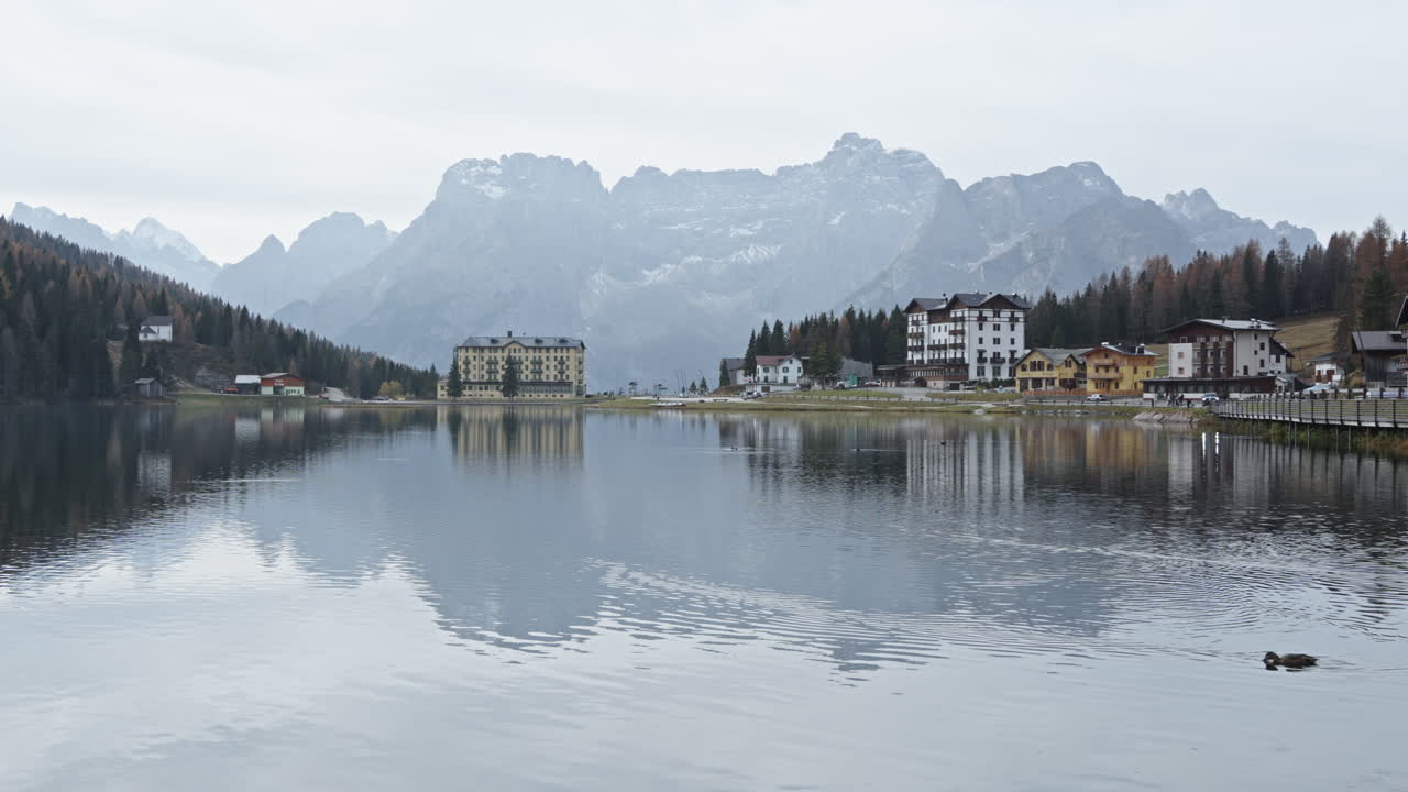 Lakeside hotels reflect in calm water with mountains in the distance on a cloudy day