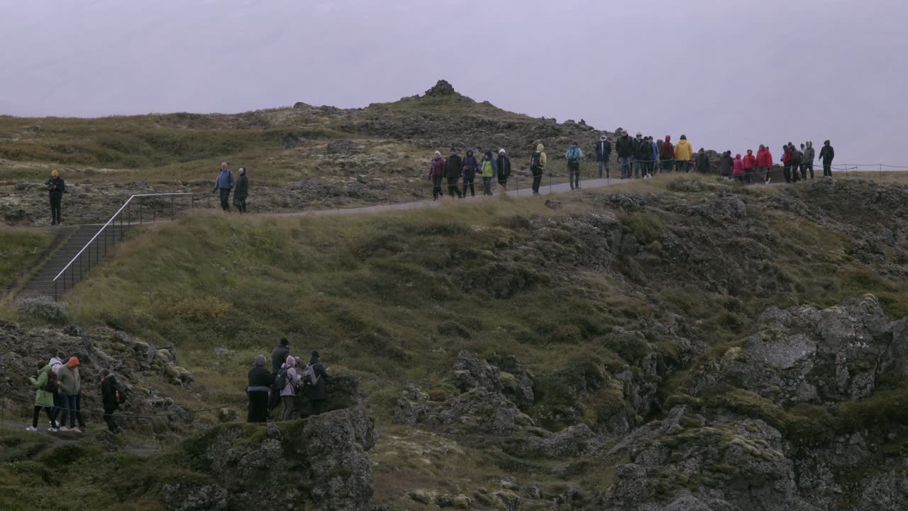 Tourists walk along a rocky cliff trail in Iceland on an overcast day with misty skies