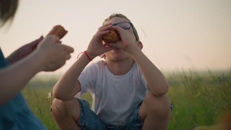 a woman in a blue dress holds two snacks as she offers one to her young son, who is wearing glasses and a white shirt. they are sitting together in a grassy field, enjoying a peaceful outdoor moment