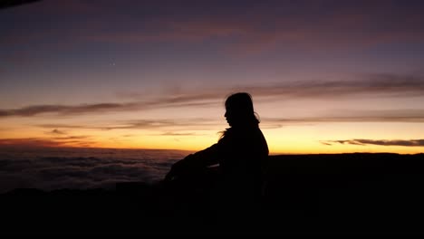 a woman sits in silence at pico do arieiro as the sunset sky glows above a cloud-filled landscape in madeira.