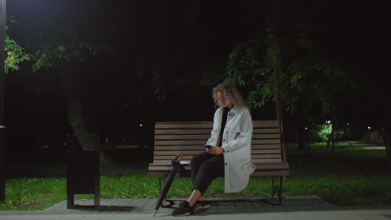 Young woman seated alone on park bench at night operating phone under bright streetlamp lifts head thoughtfully, surrounded by swaying trees and quiet green surroundings