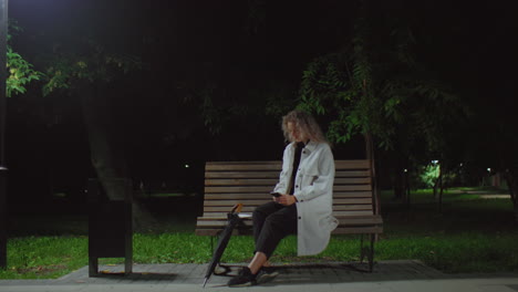 young woman seated alone on park bench at night operating phone under bright streetlamp lifts head thoughtfully, surrounded by swaying trees and quiet green surroundings