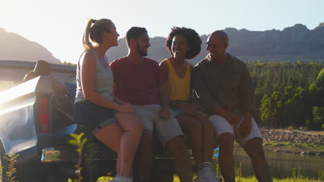 Friends-With-Backpacks-Sitting-On-Tailgate-Of-Pick-Up-Truck-On-Road-Trip-By-Lake-In-Countryside