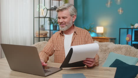 mature man sitting on table with documents and laptop smiling from successful work at home office