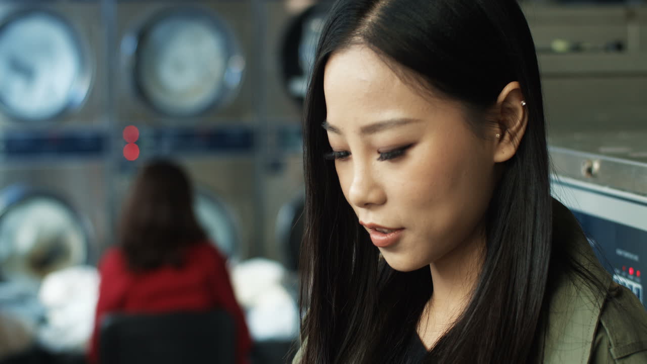 세탁실에 서 있는 동안 길고 검은 머리를 두드리고 스마트폰으로 문자 메시지를 보내는 예쁜 여성을 닫습니다