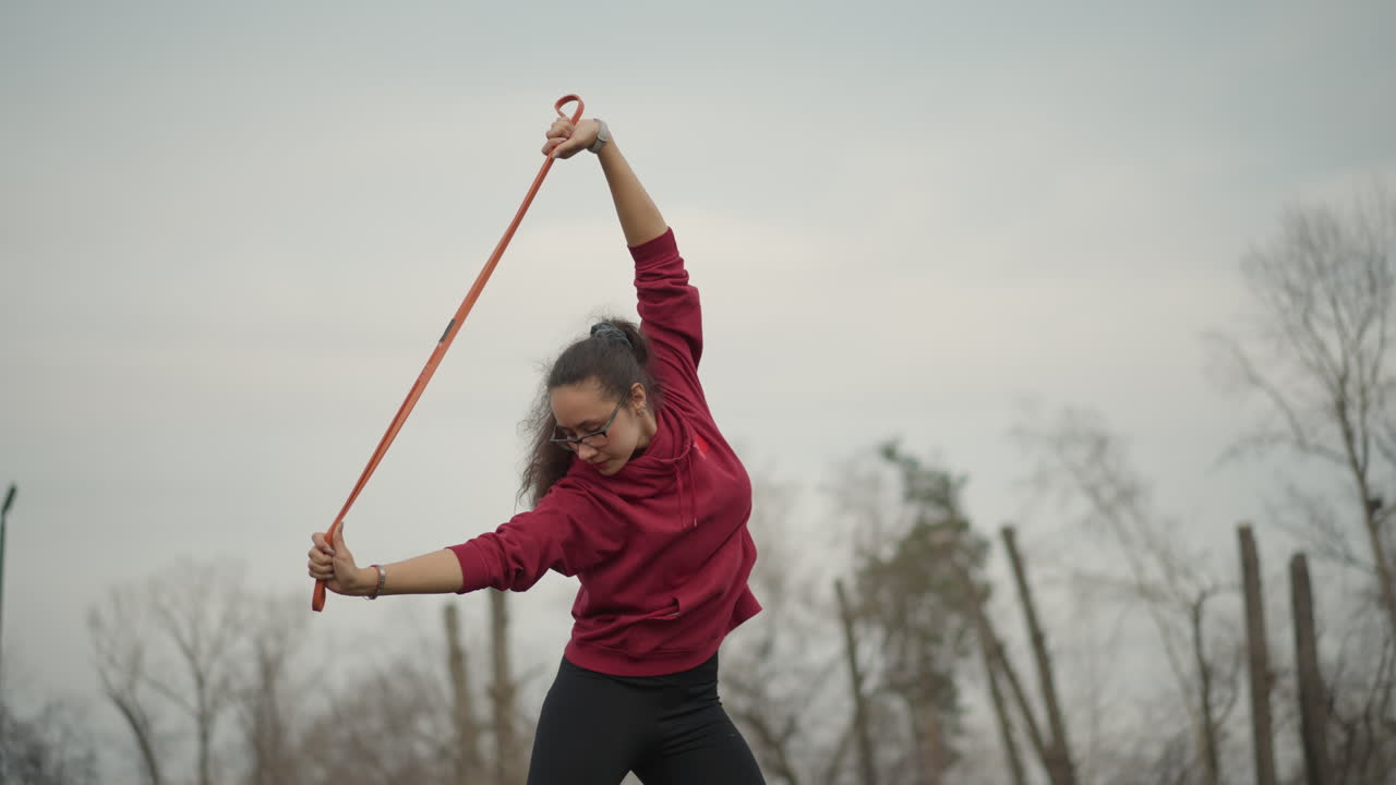 Rooftop Asian Woman Stretching With Band, Calm Morning Skyline, Hoodie Silhouette, Slow Mobility Routine Focusing On Shoulder Extension And Core Stability, Bare Trees And Soft Light Create Reflective