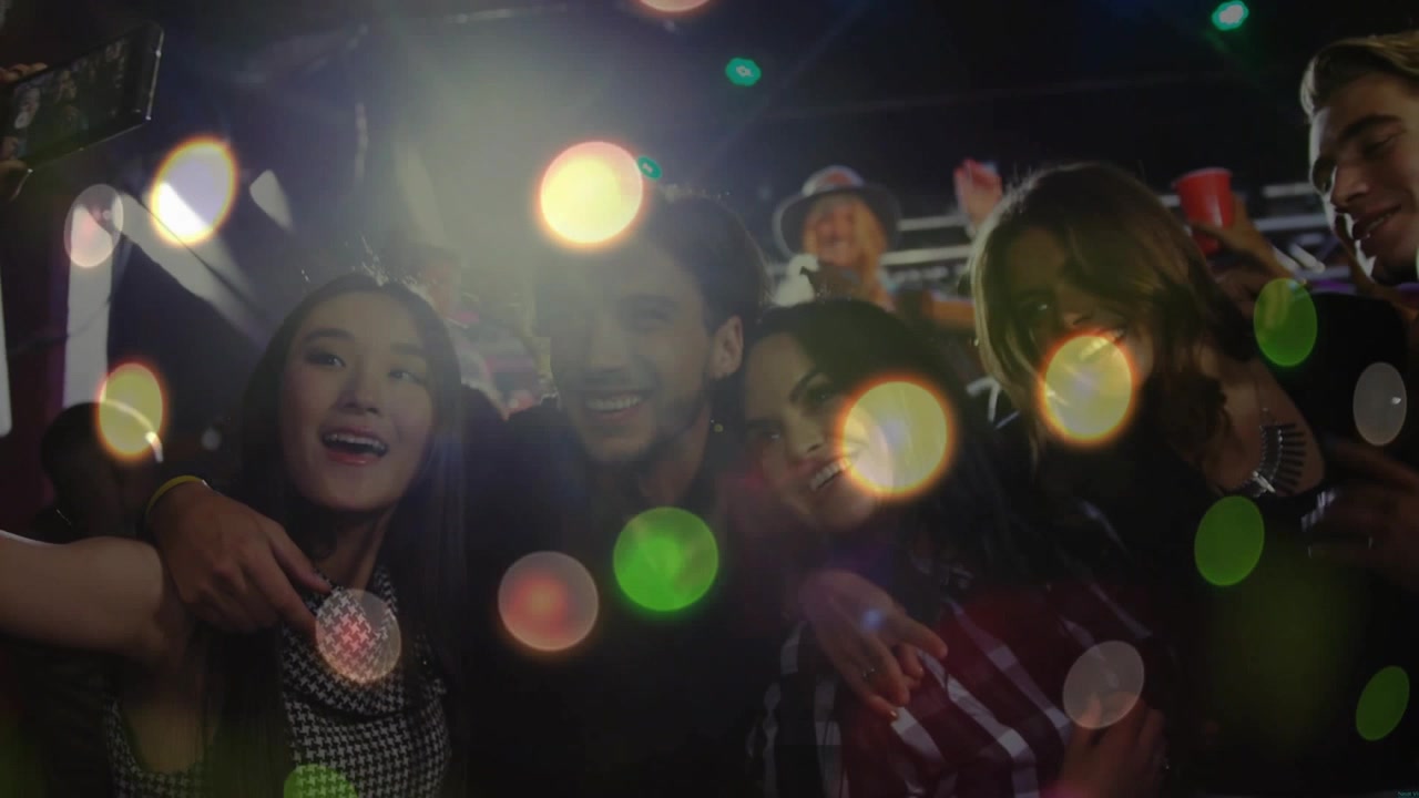Amici che si sporgono per un selfie a un concerto, donna che alza il telefono con bokeh pulsante che rivela i volti per divertimento