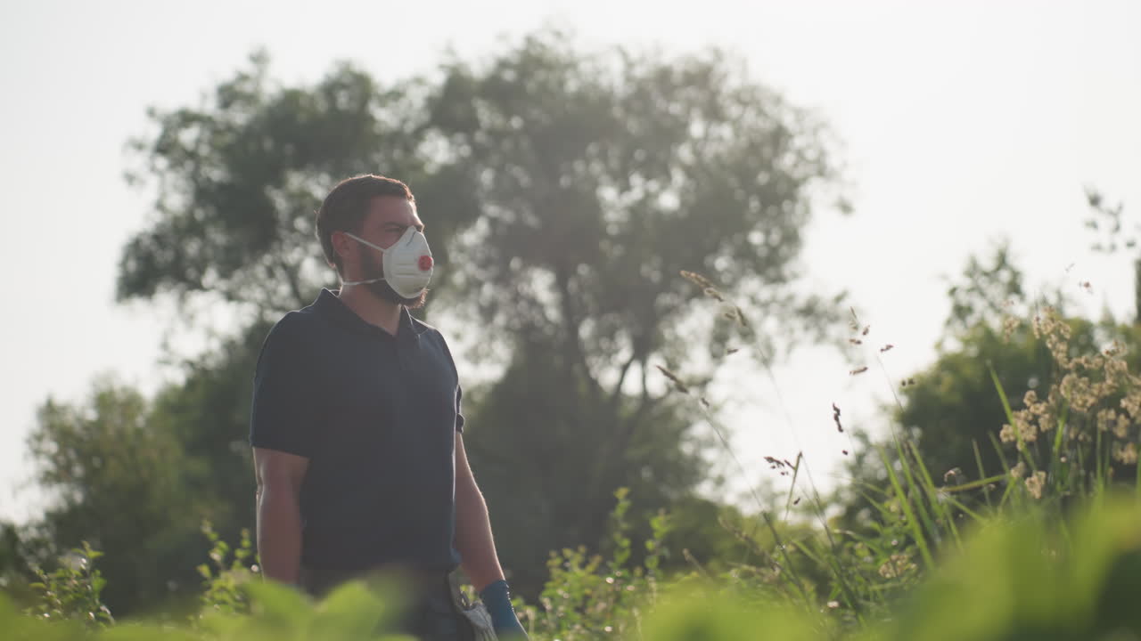 Man wearing protective mask and gloves gazes thoughtfully into distance while standing in overgrown field under sunlight, surrounded by tall grass and trees