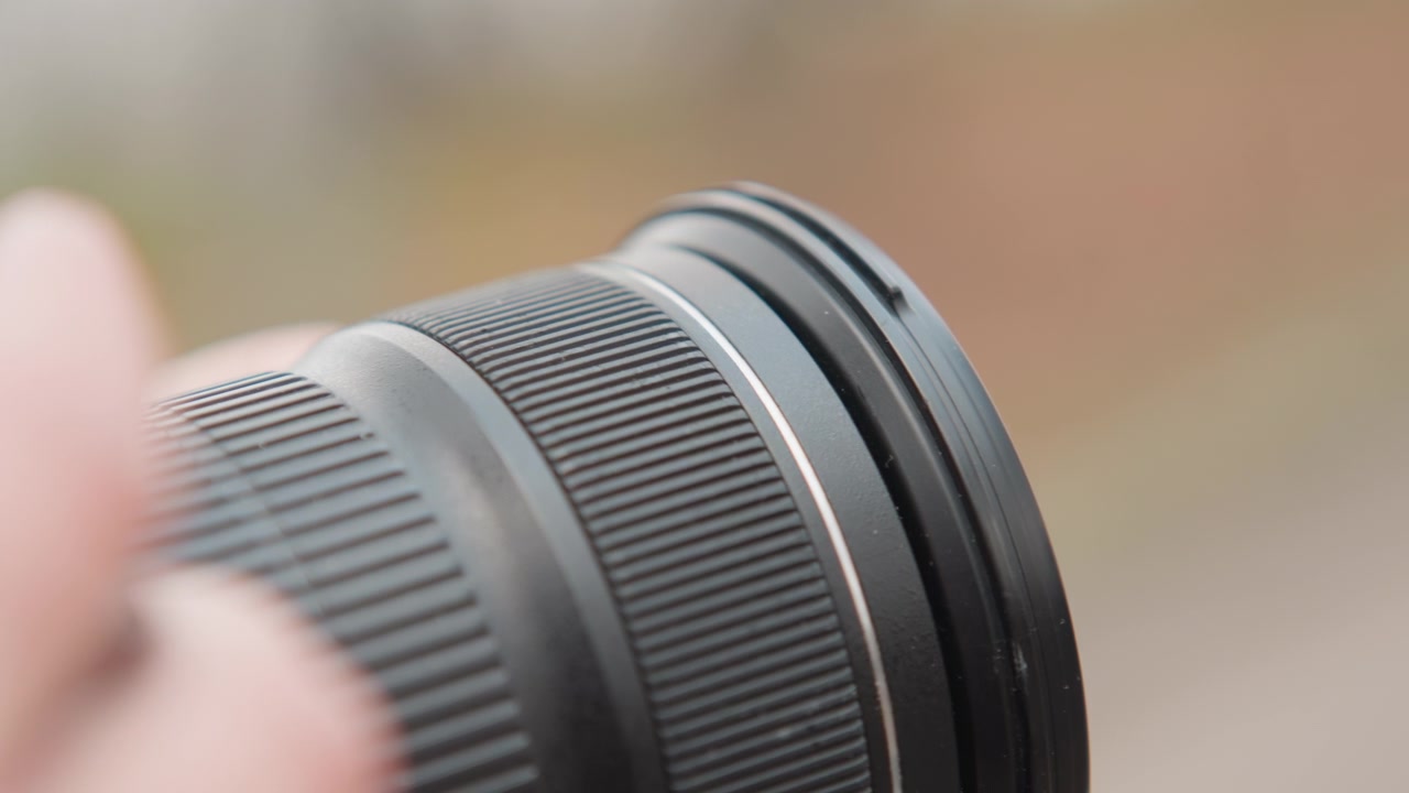 Närbild på händer som roterar objektivets fokusring. Fotografen finjusterar fokus och inspekterar fäste och textur, justerar filtergängor och testar bländaren för bokeh. Mjuk utomhusbakgrund och grunt skärpedjup.