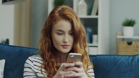 Close-Up-Of-The-Young-Beautiful-Red-Head-Girl-Sitting-On-The-Sofa-And-Typing-A-Message-Or-Scrolling-On-The-Smartphone-In-The-Living-Room