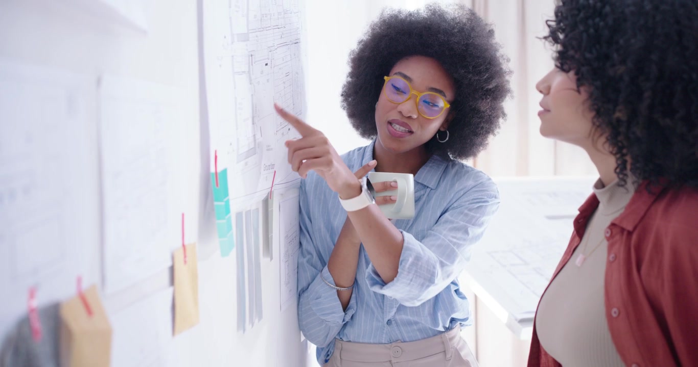 Diverse female coworkers at office plan wall, rust-red woman pointing, blue woman reviewing layout