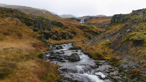 Drone-view-of-rushing-waterfalls-streaming-down-a-mountain-scape-surrounded-by-rocky-terrain-and-grasses-in-Iceland-Kirkjufell-Mountain-near-Grundarfjordour