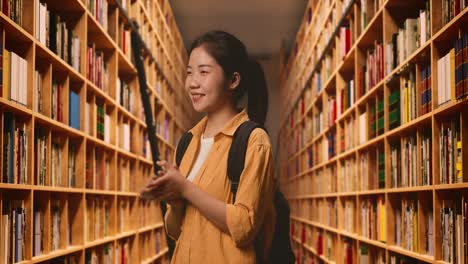 side view of asian woman student with a backpack and some books smiling and clapping her hands while standing in the library