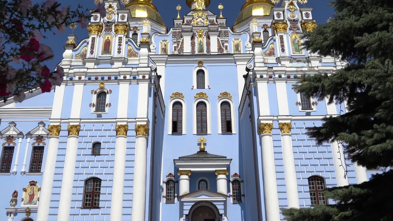Ukraine,Kyiv,St.Michael Cathedral,two young priests in black walk to the camera,some people waiting at the entrance of the iconic building painted in blue and white with golden domes and crosses,sunny