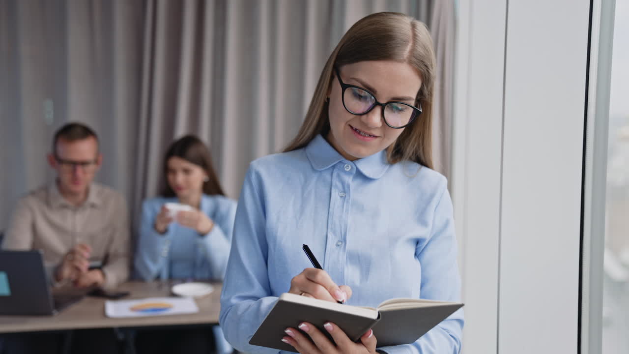 Long-haired lady in glasses taking notes into paper book standing near the window. Male and female colleagues sitting at desk at backdrop in blur.