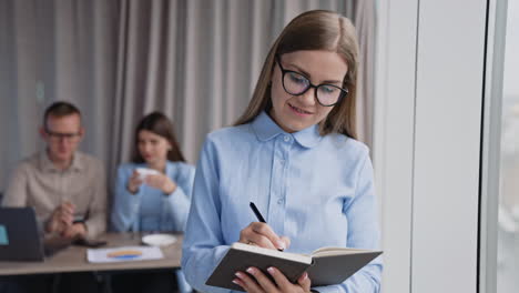 long-haired lady in glasses taking notes into paper book standing near the window. male and female colleagues sitting at desk at backdrop in blur.