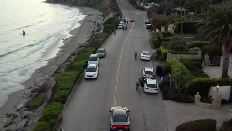 aerial drone footage flying over street with people walking next to parking cars at the ocean coast in santa barbara at sunset