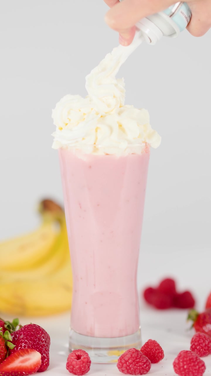 Mano che guarnisce uno smoothie rosa ai frutti di bosco con panna montata.