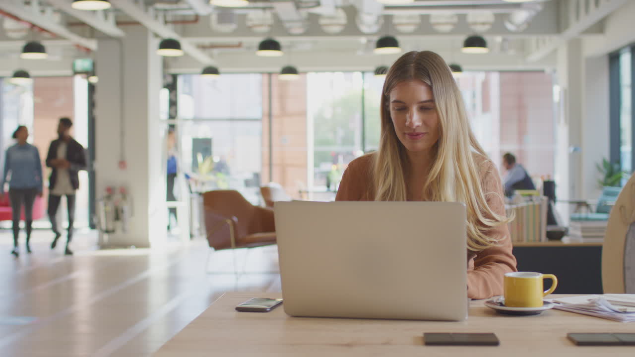 Businesswoman Working On Laptop At Desk In Modern Office With Colleagues In Background