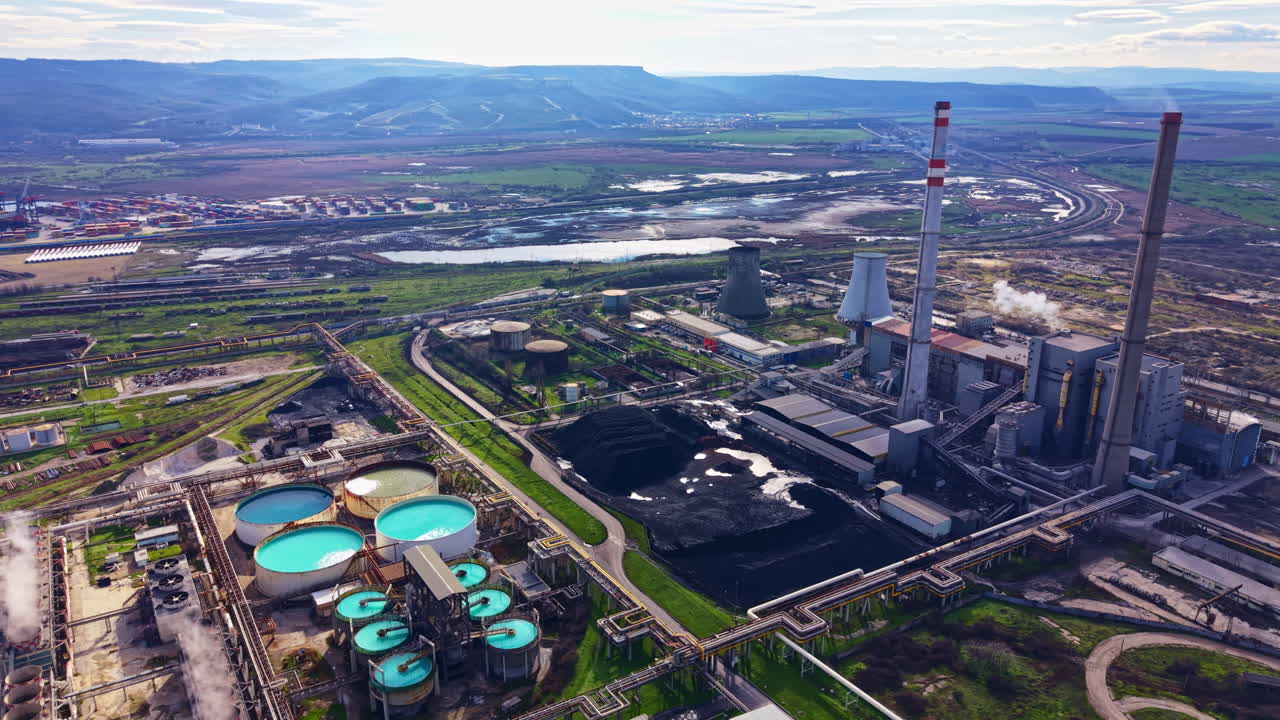 View of industrial area showing power plant, water reservoirs, and surrounding land near a river and hills during daylight