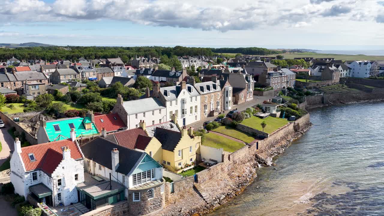 Drone glides above seaside village rooftops, historic architecture, and coastline under bright daylight