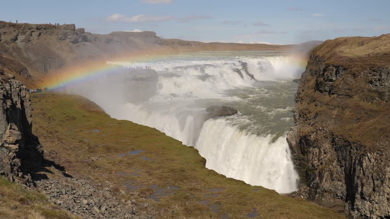 majestuosa cascada con un arco iris vibrante a través del terreno rocoso bajo un cielo despejado en islandia