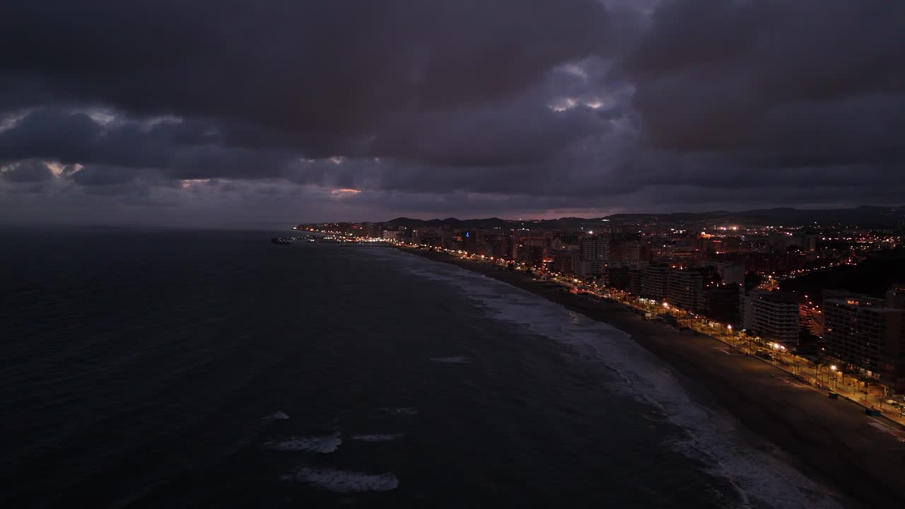 Aerial view of Torreblanca, Spain, showing the illuminated coastline at dusk under a dramatic cloudy sky, creating a moody and atmospheric scene