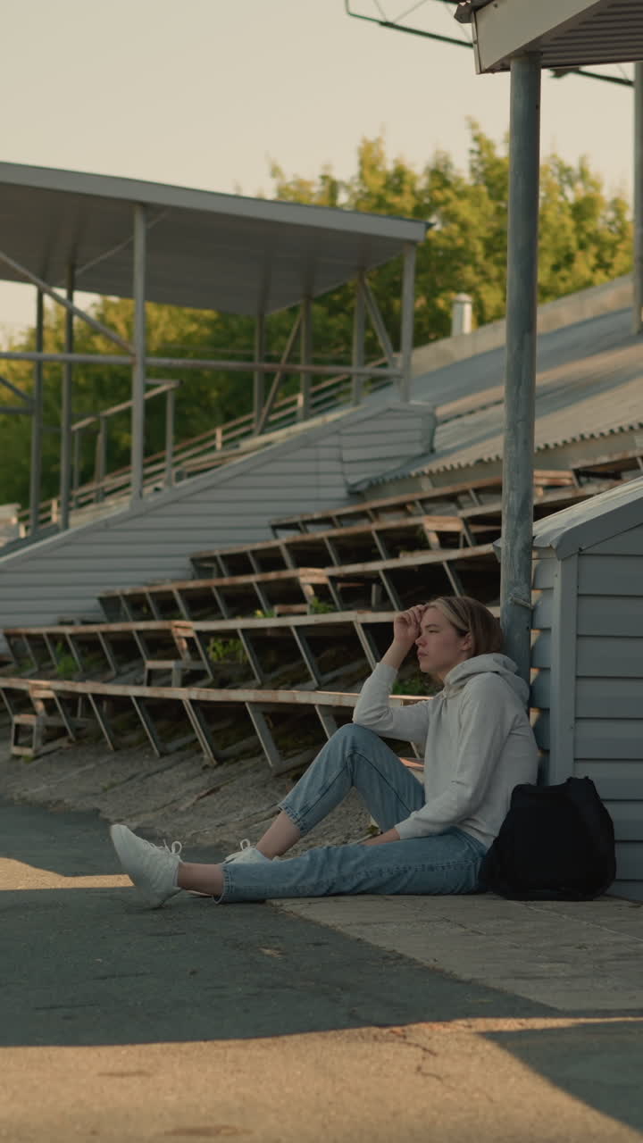 홀로 앉아있는 여인, 경기장 기둥에 기대어, 생각 많은 모습, 에 빈 관중석, 배경에는 나무, 전기 기둥, 그리고 멀리서 는 사람들의 흐릿한 그룹이 포함됩니다.