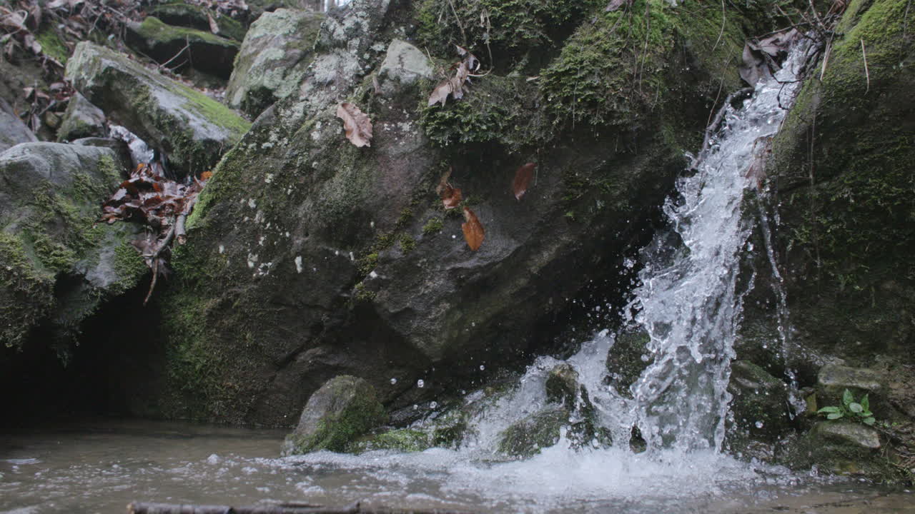 A small, picturesque waterfall cascades over moss-covered rocks into a shallow pool, surrounded by natural elements like fallen leaves and rugged stone.