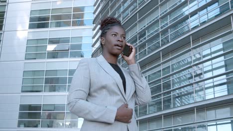 Static-shot-of-a-confident-African-American-businesswoman-standing-among-city-office-buildings-and-talking-on-mobile-phone