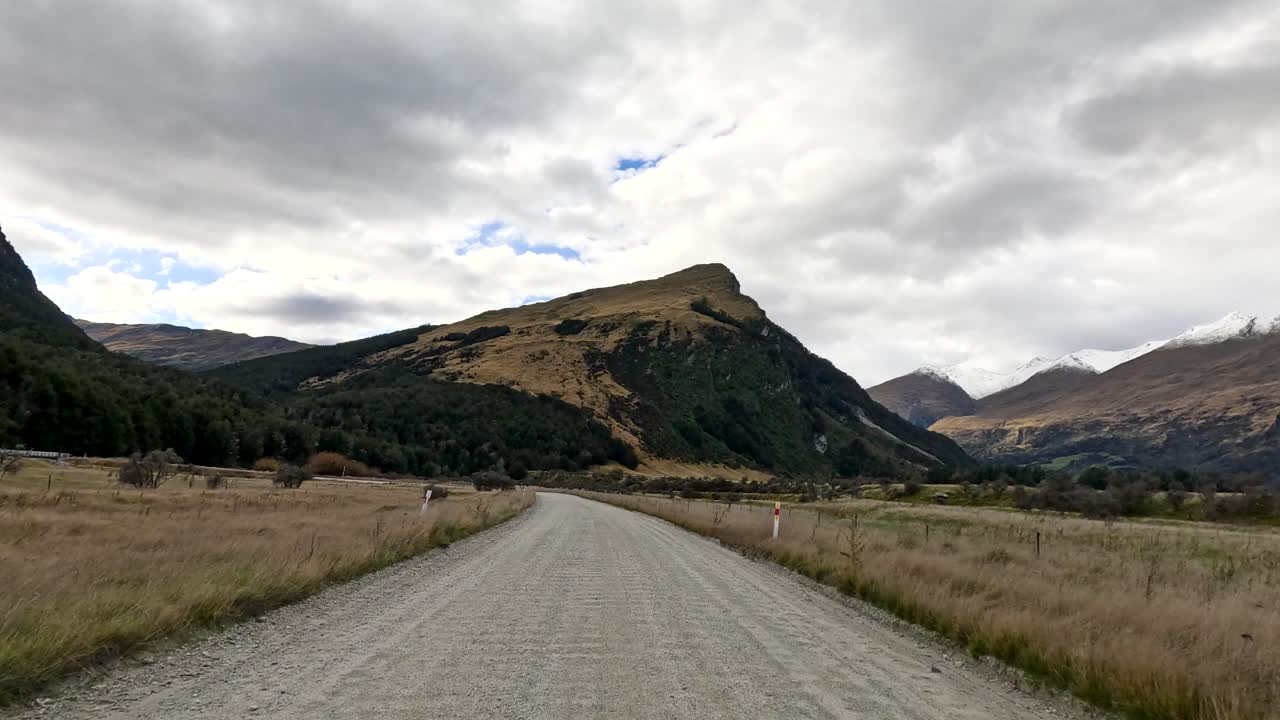 Vehicle travels along rural gravel road with overcast sky, mountains, and grassy fields in view