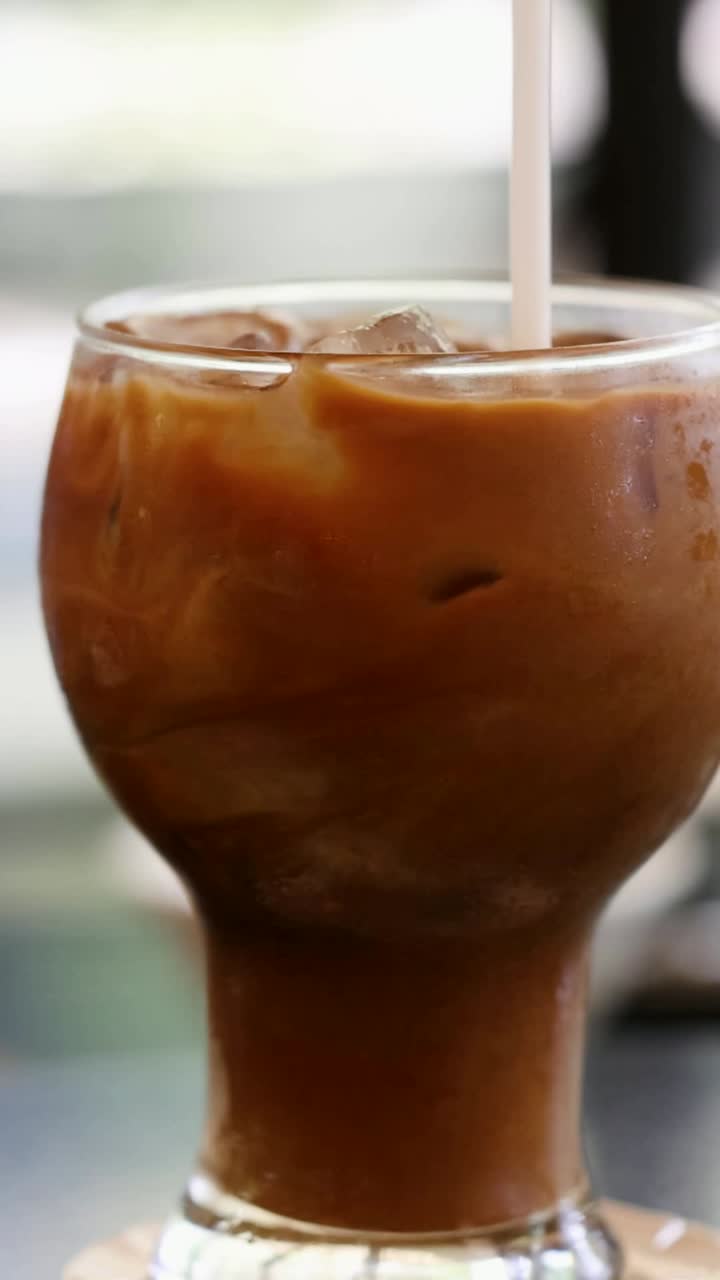 Una mano mescola un caffè freddo con una cannuccia, mettendo in risalto i motivi a spirale e la condensa in un ambiente luminoso e rilassato.
