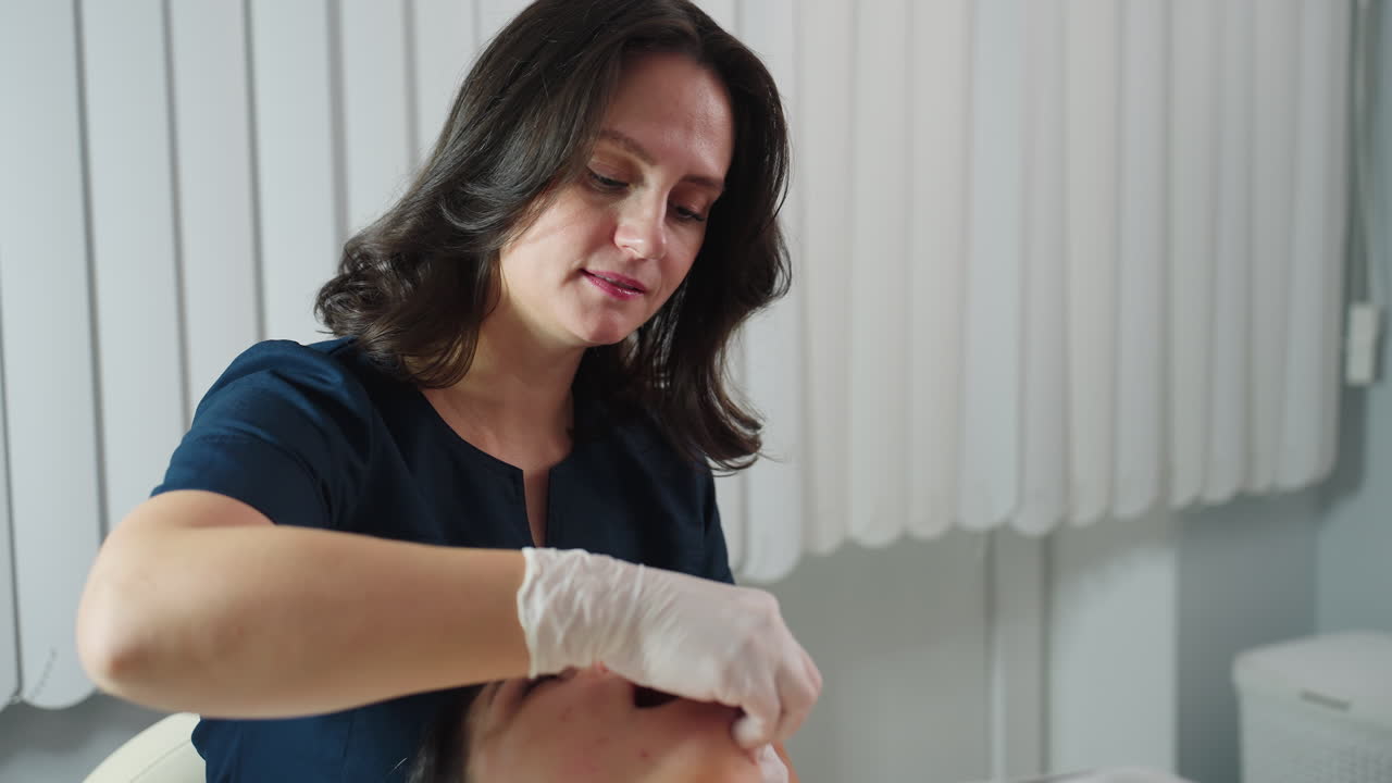 White woman practitioner wearing surgical gloves dips fingernail inside mouth of spa visitor and cradles jawline while visitor lying on massage chair closes eyes gently tenses then relaxes