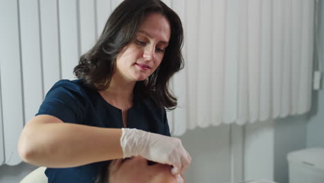 white woman practitioner wearing surgical gloves dips fingernail inside mouth of spa visitor and cradles jawline while visitor lying on massage chair closes eyes gently tenses then relaxes
