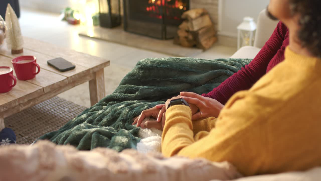 one in yellow sweater placing wrist on partner's hand, checking watch time on sofa by fireplace